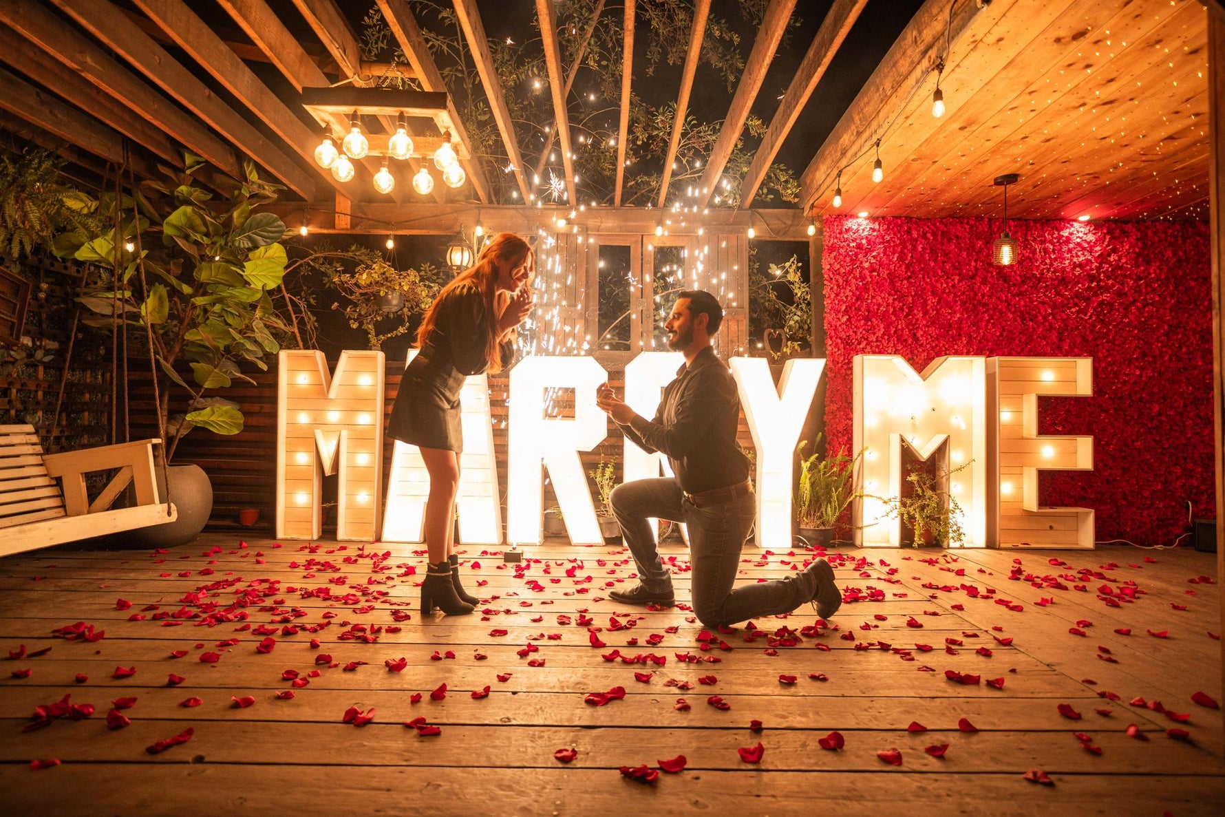 Pedida de noviazgo: él arrodillado con el anillo frente a ella, entre letras gigantes iluminadas «MARRY ME», pétalos de rosa y chispas de pirotecnia fría sobre una terraza de madera.