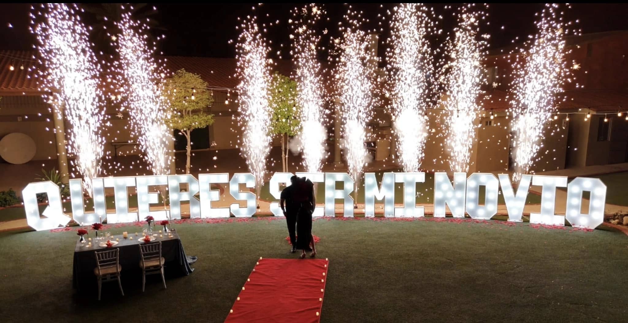 Letras luminosas de «Quieres ser mi novia» con chispas de pirotecnia fría y un pasillo de alfombra roja.