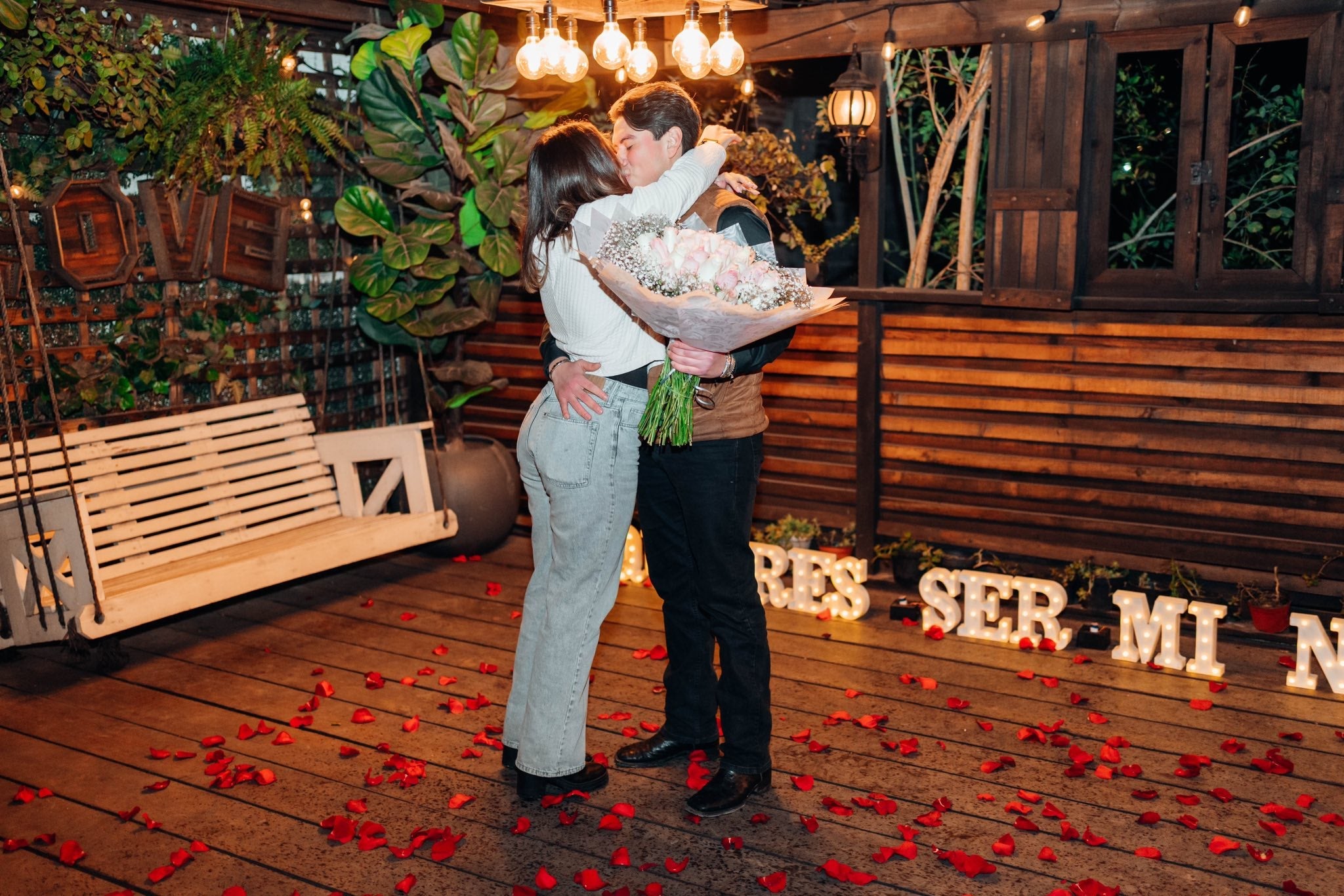 Pareja abrazándose en una terraza decorada con una mecedora blanca, pétalos de rosa en el piso y un gran ramo de rosas.